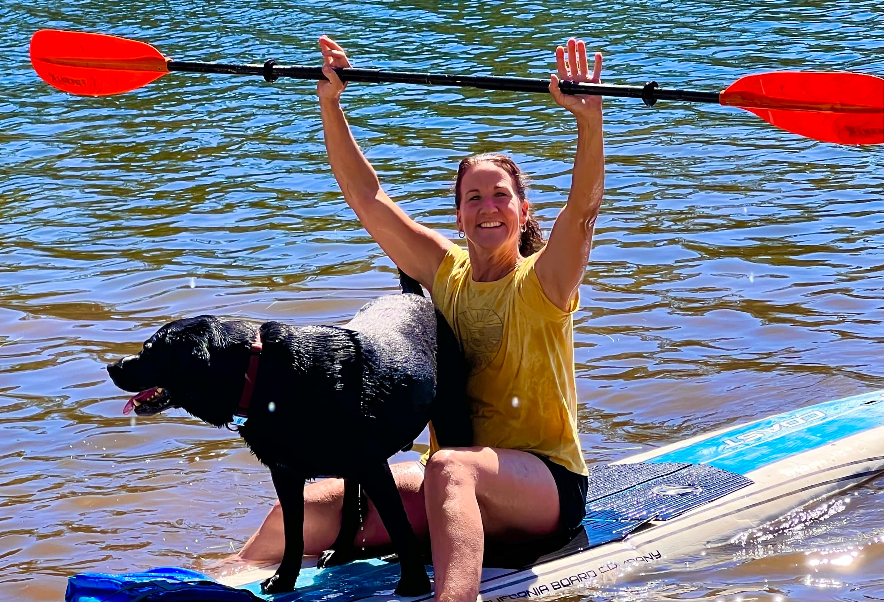 Smiling woman raises a paddle high while sitting on a paddleboard with her black dog.