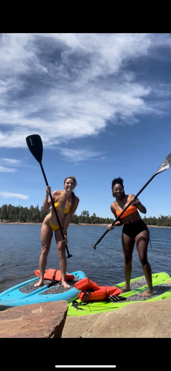 Two women smile while standing on paddleboards on a lake under blue skies.