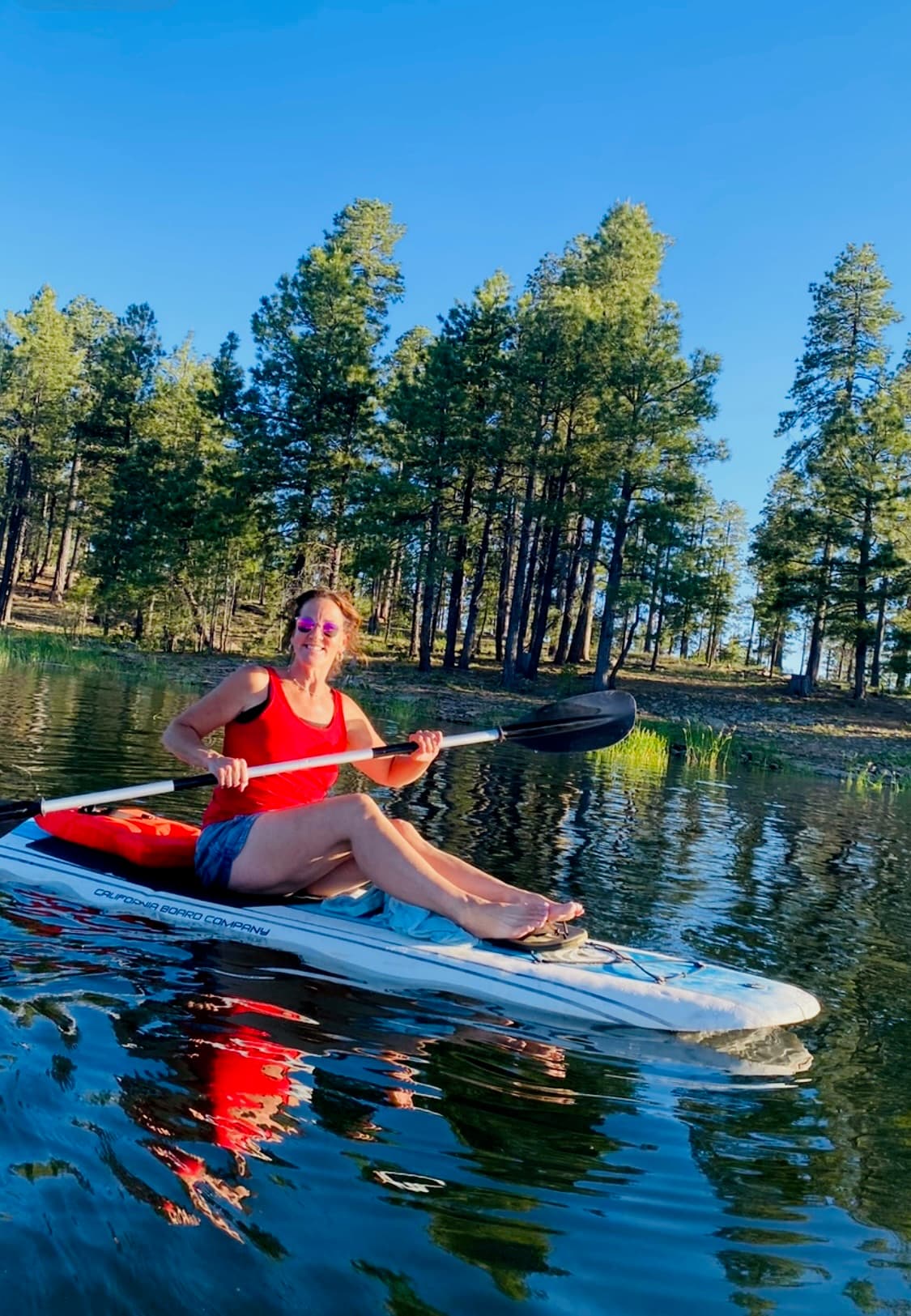 Woman in a red tank top paddles a paddleboard on a calm lake with pines.