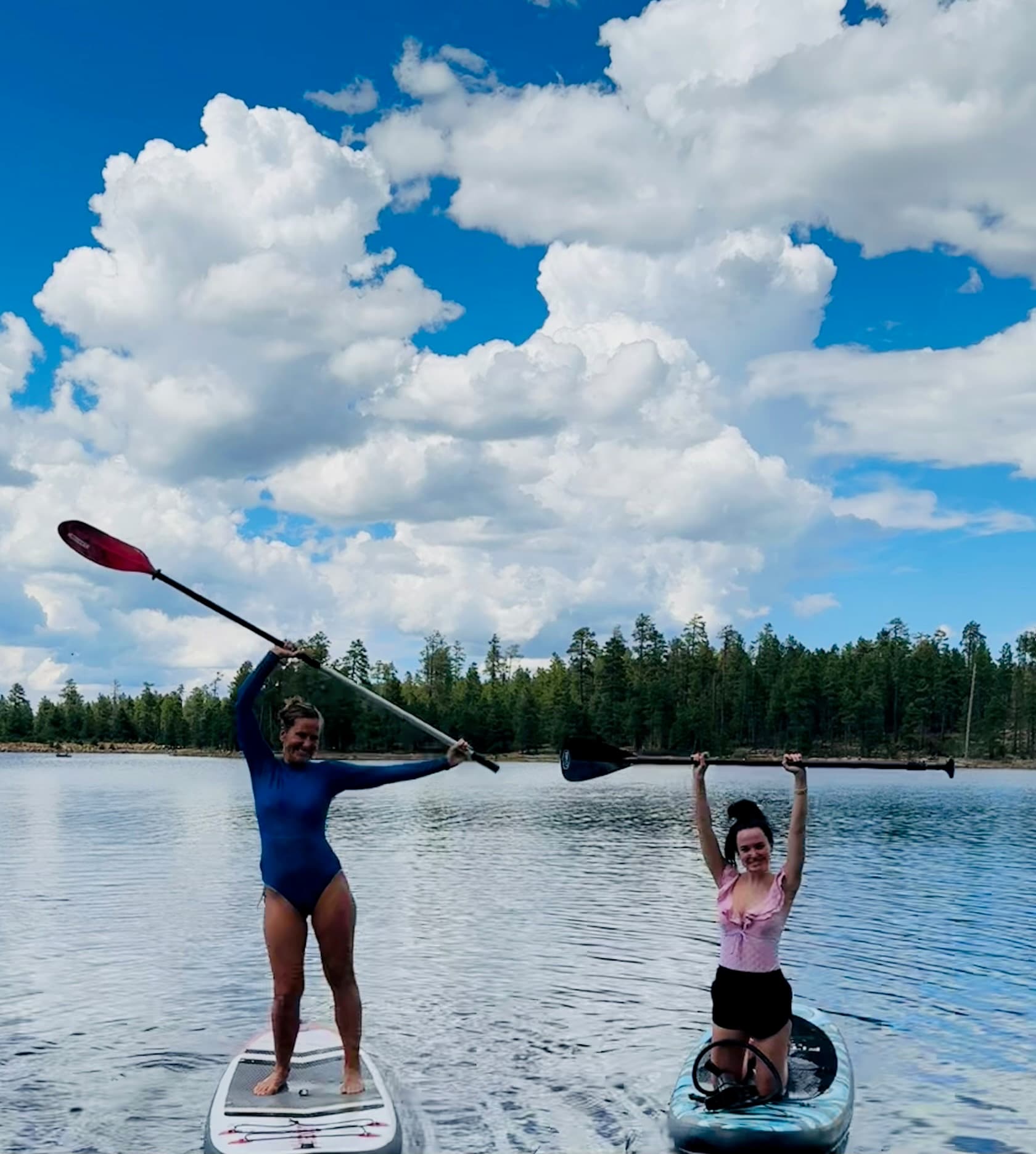 Two women pose with paddles on paddleboards and a kayak on a forest lake under massive white clouds.