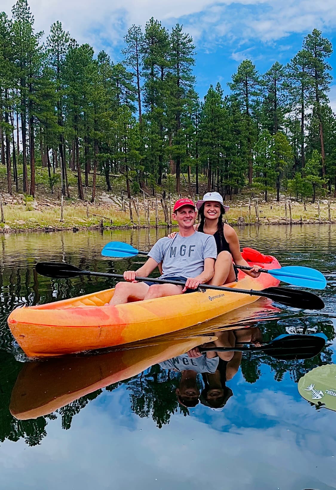 Smiling couple in an orange tandem kayak on a calm lake with a forest background.