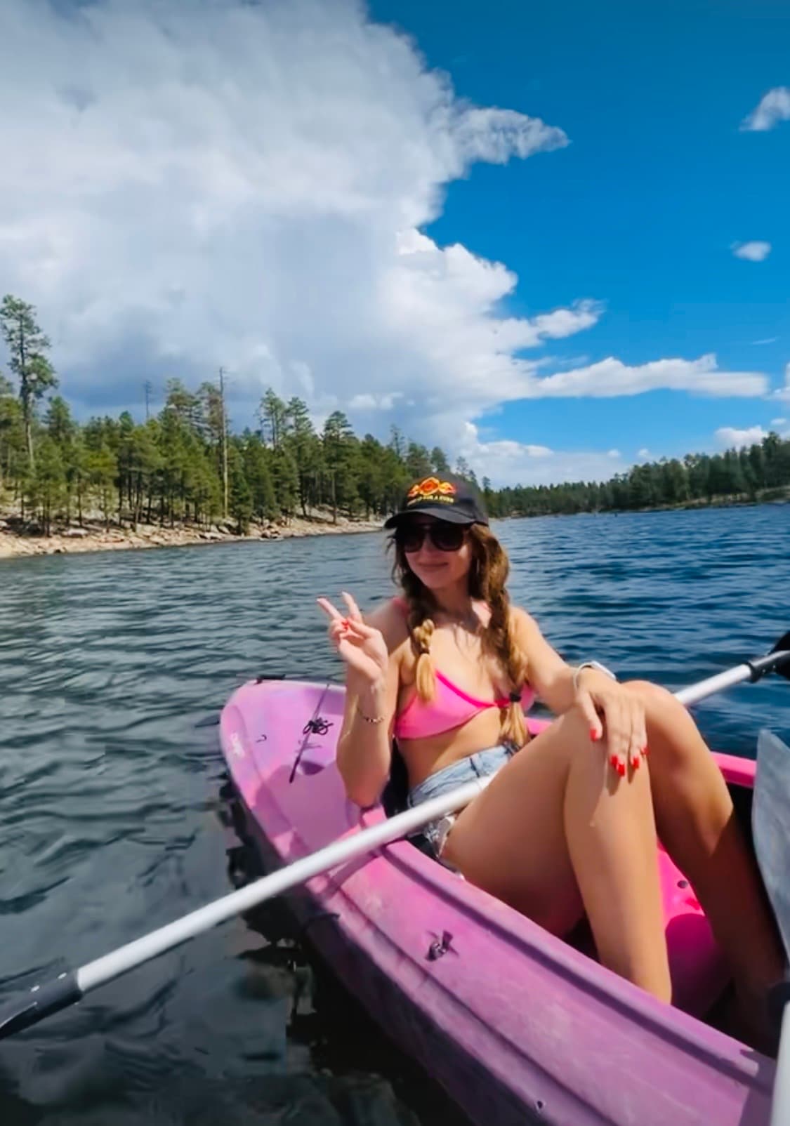 Woman in a pink kayak on a lake, wearing a bikini and making peace sign.